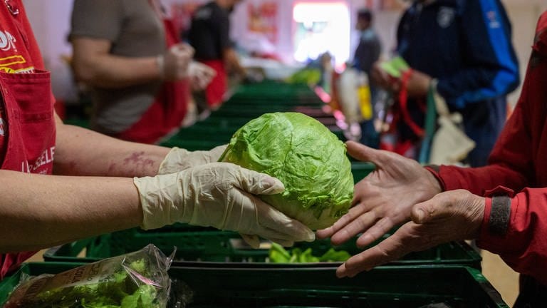 Ein Mensch überreicht einem anderen einen Salatkopf. Immer mehr Menschen in Rheinland-Pfalz sind auf Lebensmittelspenden angewiesen. (Foto: dpa Bildfunk, picture alliance/dpa | Christophe Gateau)