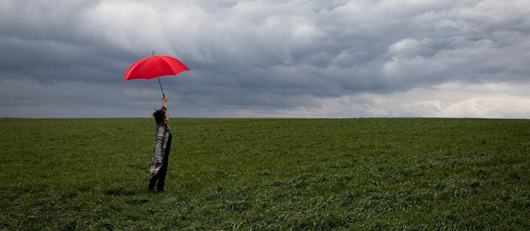 Frau mit Schirm auf Feld vor dunklem Himmel (Foto: picture-alliance / Reportdienste, picture alliance / Westend61 | FL)