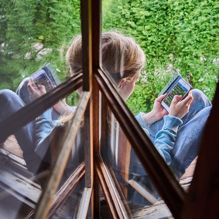 Ein Kind sitzt am Fenster und schaut auf sein Handy. (Foto: picture-alliance / Reportdienste, picture alliance/dpa | Annette Riedl)