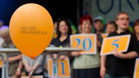 Ein Luftballon mit der Aufschrift "Tag der Organspende". Im Hintergrund Menschen, die Schilde hochalten, auf denen die Anzahl der Jahre steht, die sie nun mit Spenderorgan leben. (Foto: dpa Bildfunk, Picture Alliance)