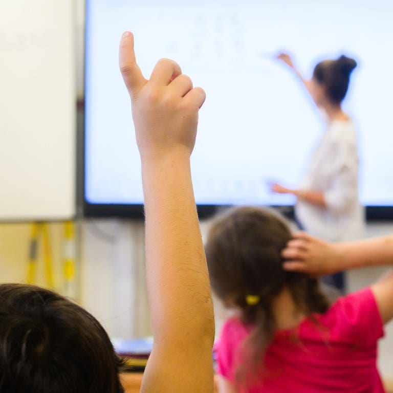 Ein Schüler meldet sich per Handzeichen, während eine Lehrerin auf eine digitale Schultafel im Klassenraum einer 4. Klasse schreibt. (Foto: dpa Bildfunk, Picture Alliance)