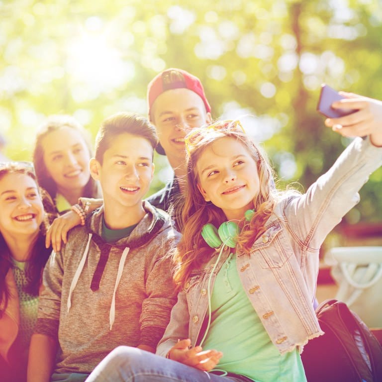 Eine Gruppe von Schülern sitzt beisammen. Sie lachen und machen ein Selfie mit dem Handy zusammen im Freien. Was in der Freizeit erlaubt ist, ist auf Klassenfahrten manchmal verboten. Denn bei manch einer Klassenfahrt muss das Handy daheim bleiben. (Foto: IMAGO, IMAGO / agefotostock)