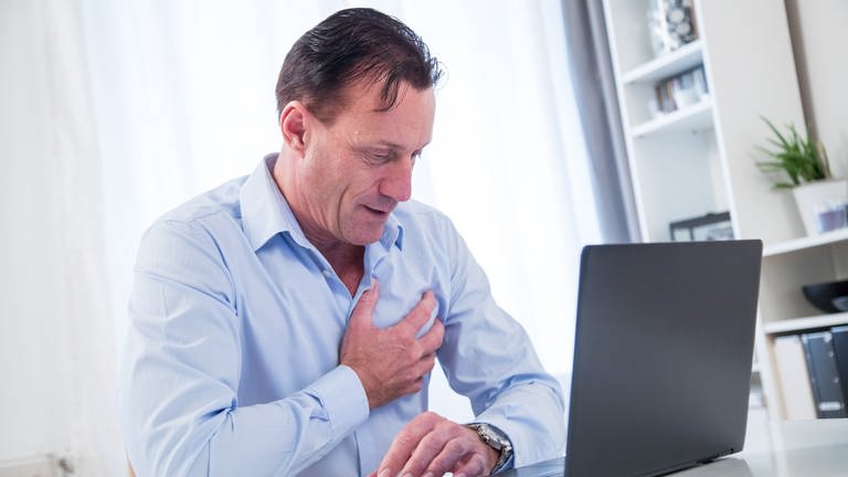 Mann sitzt am Schreibtisch vor Laptop und fasst sich mit schmerzverzerrtem Gesicht an die Brust 