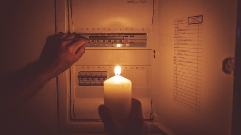 Ein Mann steht während eines Stromausfalls mit einer Kerze in der Hand vor dem Sicherungskasten.