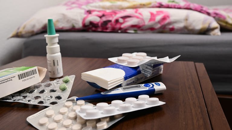 Medikamente, Taschentücher und ein Fieberthermometer liegen auf einem Nachtschränkchen neben einem Bett.