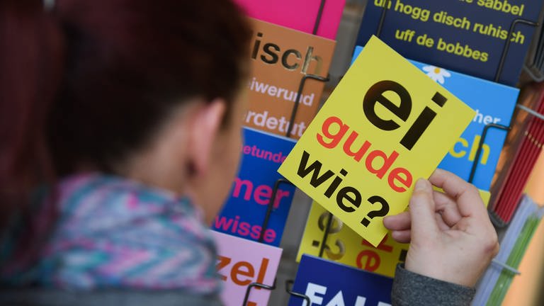 Eine Frau hält eine Postkarte mit dem klassischen hessischen Satz "Ei gude wie?" in der Hand. Jede Region hat ihre eigenen sprachlichen Einfärbungen. (Foto: dpa Bildfunk, picture alliance/dpa | Arne Dedert)