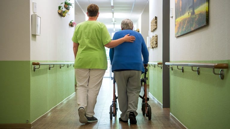 Eine Pflegefachkraft geht mit einer Bewohnerin durch ein Seniorenheim.