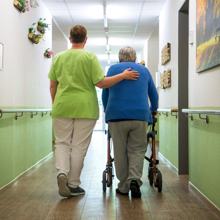 Eine Pflegefachkraft geht mit einer Bewohnerin durch ein Seniorenheim.