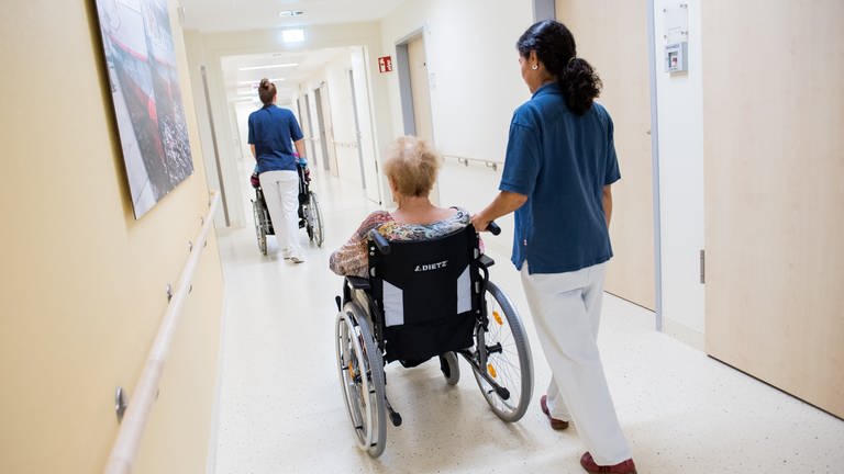 Zwei Pflegerinnen schieben Patienten mit Rollstühlen auf einem Flur in einem Krankenhaus.