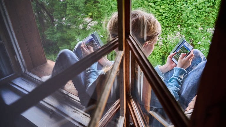 Ein Kind sitzt am Fenster und schaut auf sein Handy.