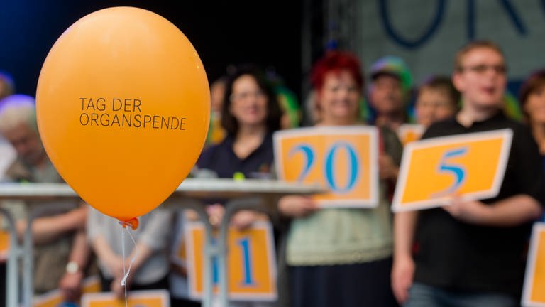 Ein Luftballon mit der Aufschrift "Tag der Organspende". Im Hintergrund Menschen, die Schilde hochalten, auf denen die Anzahl der Jahre steht, die sie nun mit Spenderorgan leben. (Foto: dpa Bildfunk, Picture Alliance)
