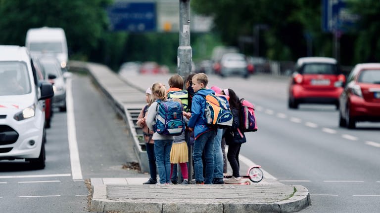 Eine Schülergruppe testet einen eventuellen, künftigen Schulweg und steht auf einer Verkehrsinsel inmitten einer viel befahrenen Straße.