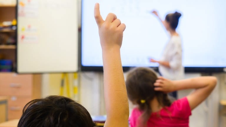 Ein Schüler meldet sich per Handzeichen, während eine Lehrerin auf eine digitale Schultafel im Klassenraum einer 4. Klasse schreibt.