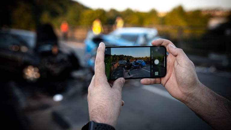 Ein Gaffer macht mit seinem Smartphone Bilder von einer Unfallstelle. In Mainz wurden nach einem tödlichen Unfall Bilder und Videos von einem der Opfer gemacht und ins Netz gesetellt. Die Polizei ermittelt.