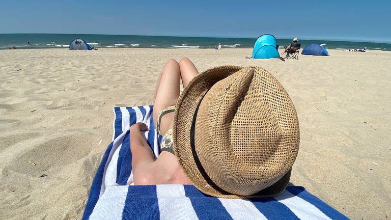 Sommer, Urlaub, Ferien: Eine Urlauberin mit Sonnenhut liegt am Sandstrand auf einem Handtuch und schaut auf das Meer. (Foto: picture-alliance / Reportdienste, picture alliance / Kirchner-Media | David Inderlied)