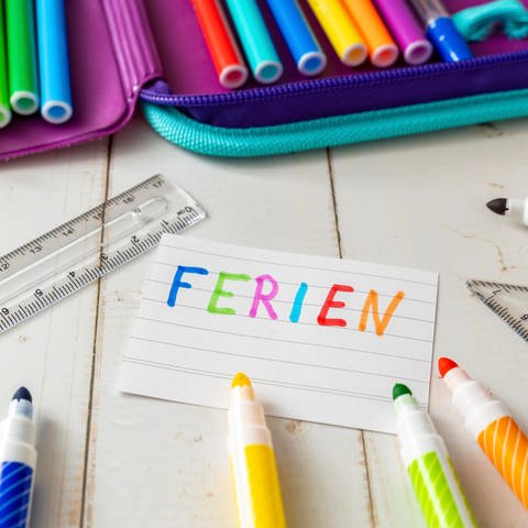 Symbolbild: "Ferien" als Notiz mit bunter Farbe geschrieben, neben einem Schulmäppchen und bunten Stiften