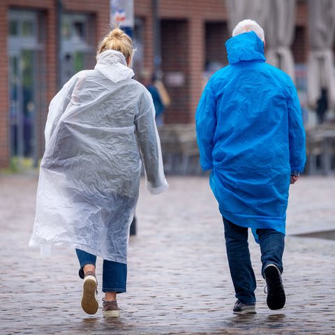 Ein Mann und eine Frau laufen in Regenponchos über eine nasse gepflasterte Straße - es stürmt.