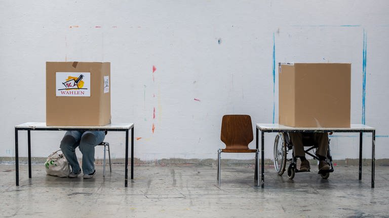Wahlberechtigte sitzen einem Wahllokal in Trier hinter einem Sichtschutz, der als Wahlkabine gilt, um ihre Stimme abzugeben. In vielen Gemeinden in RLP fehlen bei den anstehenden Kommunalwahlen Kandidaten für das Ortsbürgermeisteramt. 