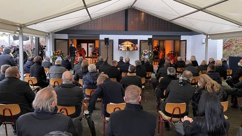 Etwa 50 Gäste sitzen in einem Zelt vor der Trauerhalle, in der die Trauerfeier für den verstorbenen Horst Eckel stattfindet. (Foto: SWR)
