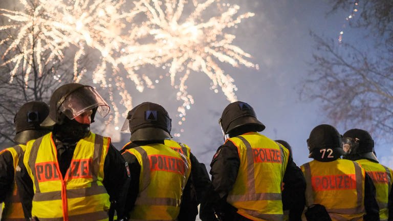 Mehrere Polizisten mit Warnwesten und Helmen stehen im Vordergrund. Im Hintergrund ist Feuerwerk zu sehen.  (Foto: dpa Bildfunk, picture alliance/dpa | Clemens Heidrich)