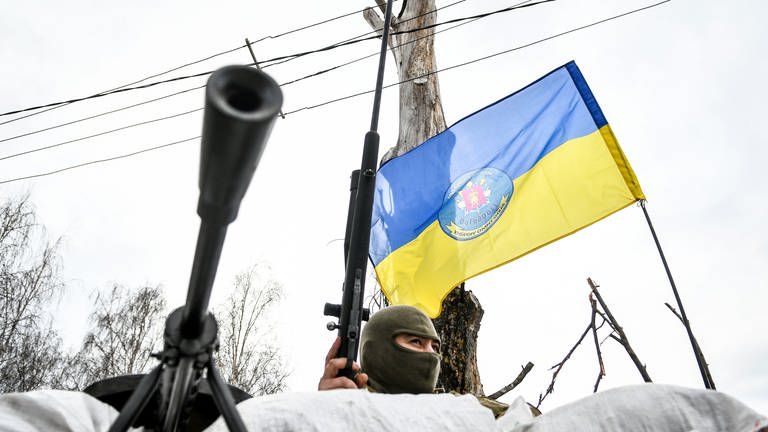 “Russland-Ukraine-Konflikt: Ein Soldat aus der Ukraine sitzt hinter Sandsäcken. Er trägt eine Sturmhaube und hat ein Gewehr in der Hand. Im Hintergrund weht die Ukraine-Flagge. (Foto: dpa Bildfunk, picture alliance/dpa/Ukrinform )