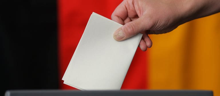 Eine Hand wirft einen Stimmzettel in eine Urne - dahinter ist die Deutschlandfahne. (Foto: dpa Bildfunk, picture alliance / dpa | Jens Wolf)