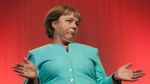 Marina Tamássy steht als Angela Merkel auf der Bühne. Sie trägt eine grüne Jacke.  (Foto: Pressestelle, Die Untiere)