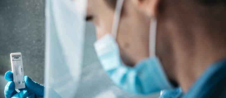Eine Frau mit Mundschutz hält einen Corona-Test in der Hand (Foto: IMAGO, IMAGO / Bihlmayerfotografie)