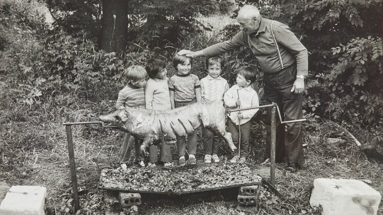 Schwarz-Weiß-Foto mit älterem Mann, der einer Gruppe Kinder vor einem Spanferkel die Köpfe tätschelt (Foto: SWR, Fotograf: Fabio Sgroi)