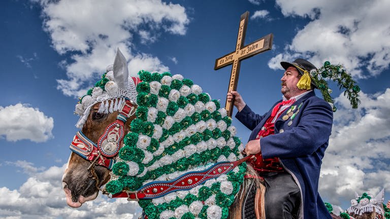 Mann mit Holzkreuz in der Hand sitzt bei einer Prozession auf einem geschmückten Pferd (Foto: SWR, Fotograf: Hans-Jürgen Burkard)