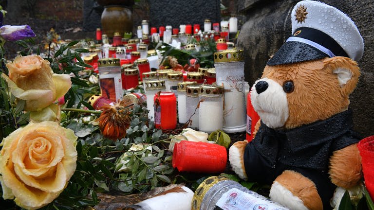 Ein Teddybär im Polizistenkostüm liegt zwischen Trauerbekundungen, Blumen und Kerzen. (Foto: IMAGO / BeckerBredel)