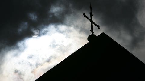 Im Gegenlicht und vor wolkenverhangenem Himmel ist die Kirchturmspitze eines Doms mit Kreuz zu sehen. (Foto: picture-alliance / Reportdienste, picture alliance/dpa | Friso Gentsch)
