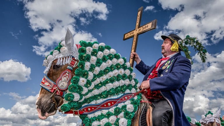 Mann mit Holzkreuz in der Hand sitzt bei einer Prozession auf einem geschmückten Pferd (Foto: SWR, Fotograf: Hans-Jürgen Burkard)