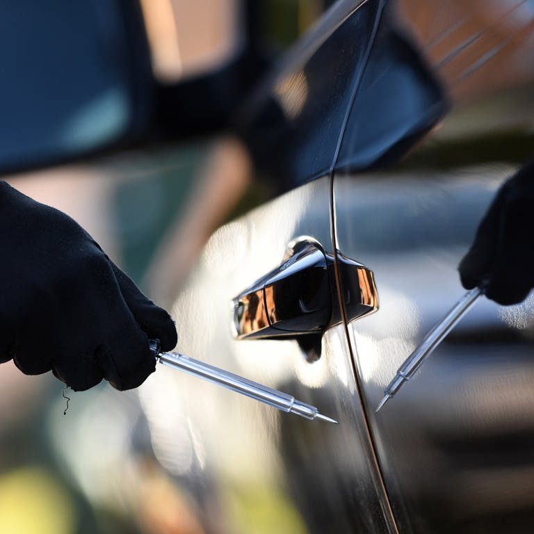 Ein Mensch mit einem schwarzen Handschuh zerkratzt mit einem spitzen Gegenstand eine Autotür. 