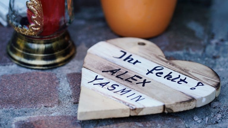 Auf einem Holzbrett in Herzform ist an einer Landstraße zwischen Kusel und Ulmet an einer Gedenkstätte für zwei getötete Polizisten die Aufschrift „Ihr fehlt! Alex Yasmin“ aufgebracht.