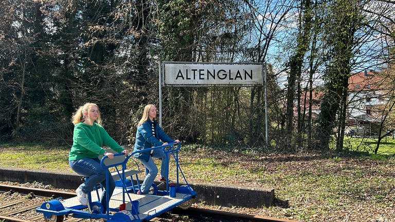 Zwei junge Frauen fahren auf einer Draisine durch die Landschaft, hinter ihnen das Ortsschild von Altenglan