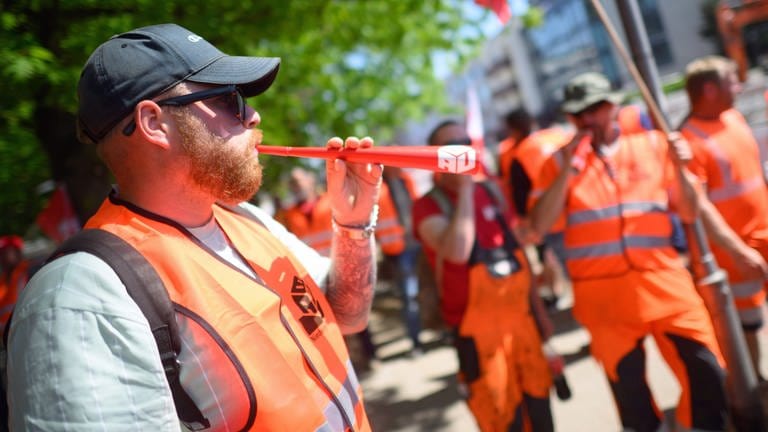 Bauarbeiter hält bei Demonstration eine Tröte in der Hand und bläst hinein - IG Bau streikt auch im Westen der Pfalz
