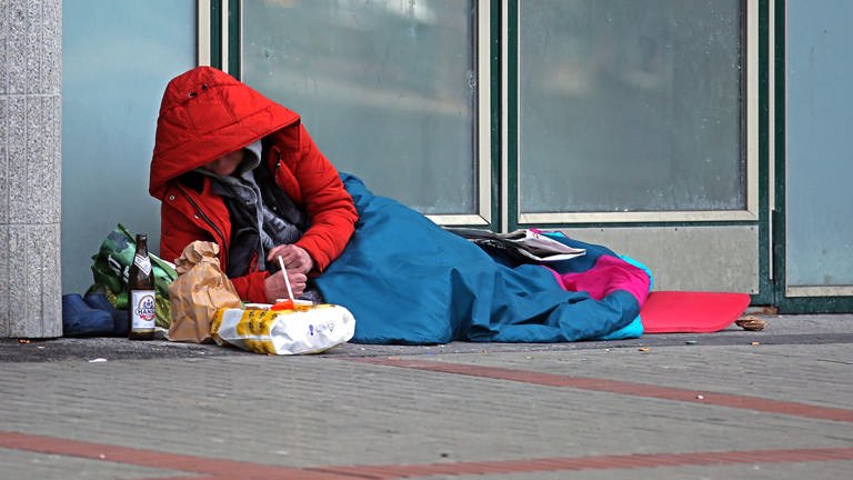 Ein Mensch liegt auf dem Boden, eingepackt in eine dicke Jacke und einen Schlafsack.In Koblenz und der Region bieten viele Vereine und Organisationen Wohnungslosen auch in diesem Winter warmes Essen und Kleidung, Übernachtungsplätze und Schlafsäcke an.  (Foto: IMAGO, imago images/Gottfried Czepluch)