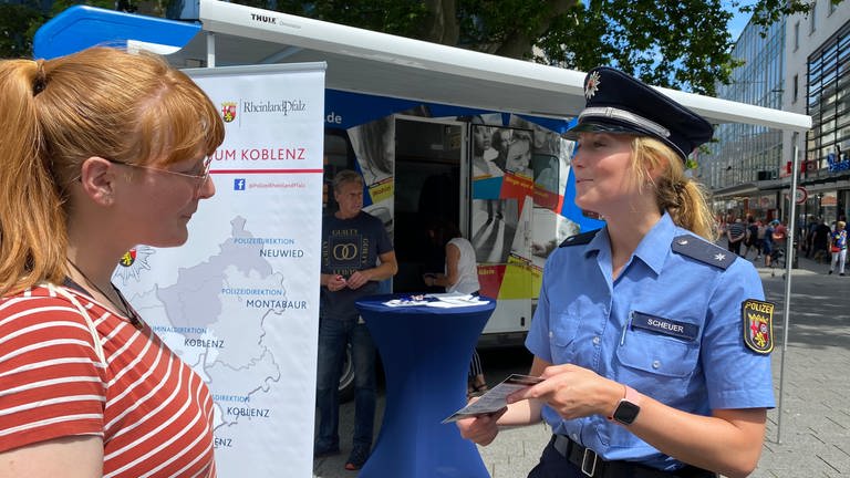 Eine Polizistin überreicht in der Fußgängerzone in Koblenz an einem Infostand einen Flyer an eine Fußgängerin. (Foto: SWR)