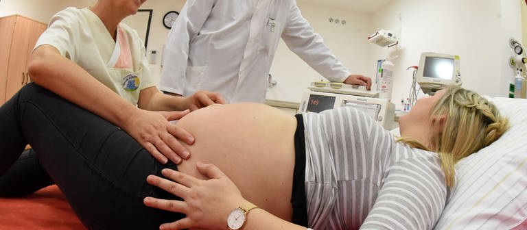 Eine schwangere Frau liegt im Krankenhaus auf einer Liege und wird von einer Hebamme und einem Arzt untersucht (Foto: picture-alliance / Reportdienste, picture alliance / Waltraud Grubitzsch/dpa-Zentralbild/dpa)