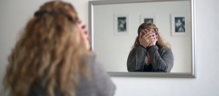 Eine Frau steht vorm Spiegel und hält sich verzweifelt die Hände vors Gesicht, sie ist nicht zu erkennen. (Foto: picture-alliance / Reportdienste, picture alliance / dpa | Ina Fassbender)