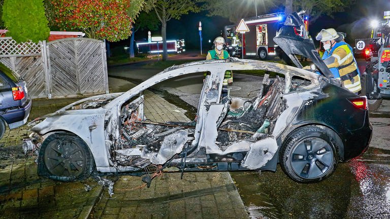 Ein ausgebranntes E-Auto, die Feuerwehr steht dabei (Foto: IMAGO, IMAGO / KS-Images.de)