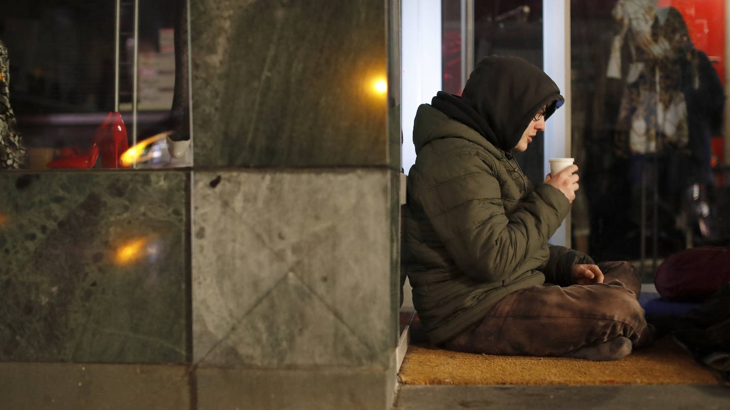 Ein obdachloser, junger Mann sitzt an einer Mauer einer Hauswand eines Geschäfts und trinkt aus einem Becher (Foto: picture-alliance / Reportdienste, biky)