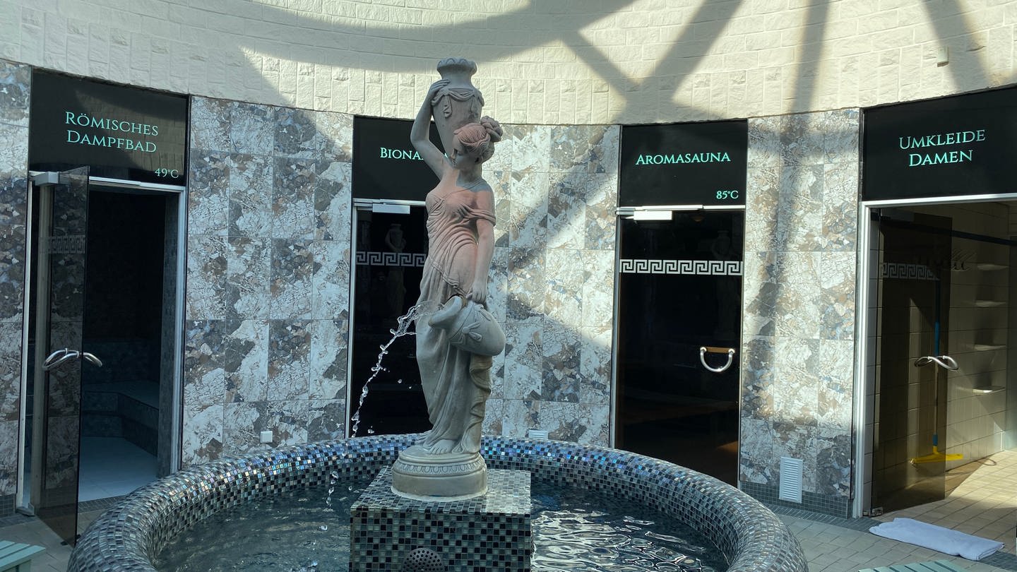 Ein Brunnen mit einer weiblichen Statue, die Amphoren trägt. Dahinter sieht man die Türen der verscheidenen Saunen und der Umkleiden. (Foto: SWR)