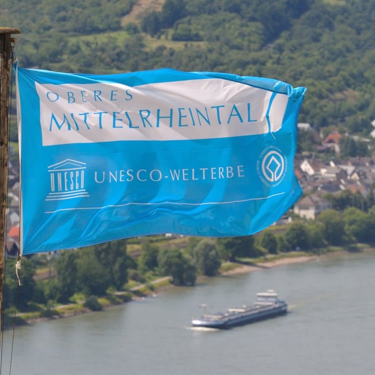 Eine Fahne mit der Aufschrift "Oberes Mittelrheintal - UNESCO-Welterbe" weht am 27.07.2009 über der Marksburg in Braubach bei Koblenz, während im Hintergrund ein Frachtschiff auf dem Rhein zu sehen ist.