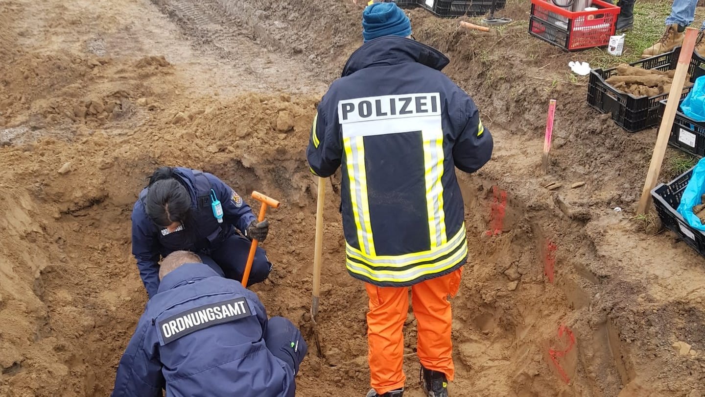 Drei Menschen in Arbeitskleidung stehen und hocken mit Schaufeln in einer Grube. Zwei von ihnen tragen Jacken mit der Beschriftung 