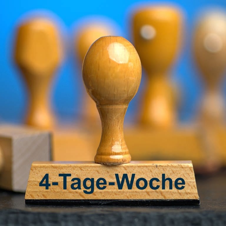 Ein Holzstempel mit der Aufschrift "4-Tage-Woche" auf einer Schieferplatte mit weiteren Stempeln unscharf dahinter. In Koblenz und Bad Neuenahr machen zwei Handwerksbetriebe mit der Vier-Tage-Woche gute Erfahrungen