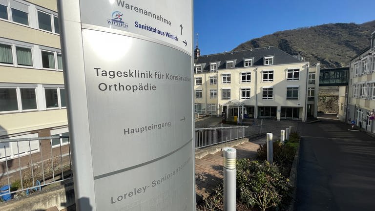 Schild mit Aufschrift Tagesklinik für Konservative Orthopädie. Im Hintergrund die Klinik und das Mittelrheintal in Oberwesel.