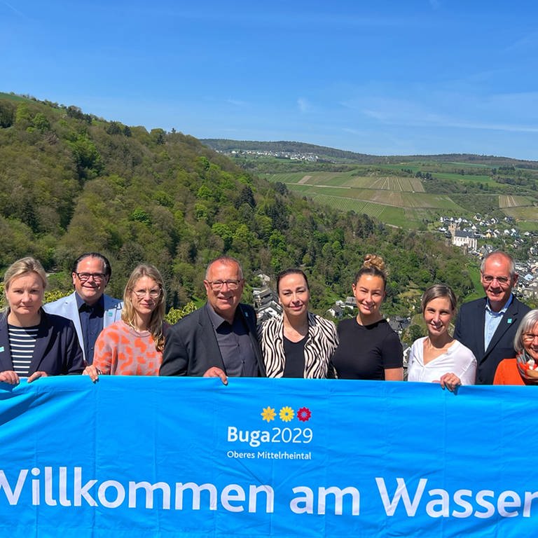 Verantwortliche der BUGA 2029 am Mittelrhein halten ein blaues Banner mit der Aufschrift "Willkommen am Wasser".  (Foto: Bundesgartenschau Oberes Mittelrheintal 2029 gGmbH)