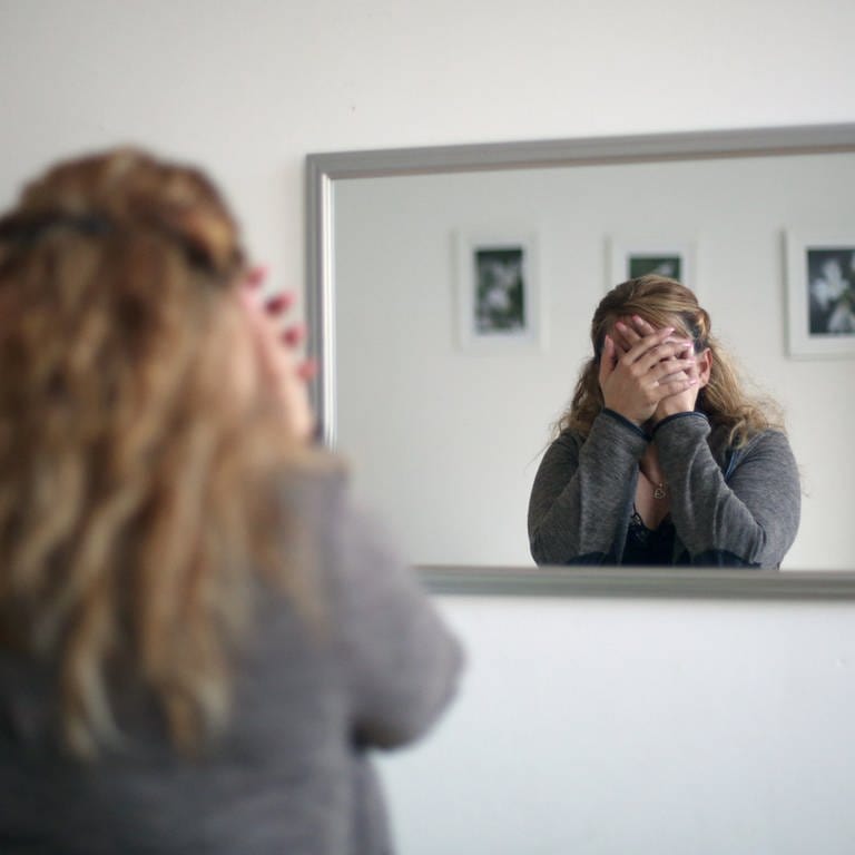 Eine Frau steht vorm Spiegel und hält sich verzweifelt die Hände vors Gesicht, sie ist nicht zu erkennen.